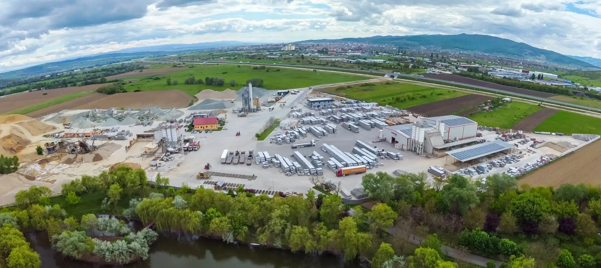 Vedere aeriană a unei platforme de producție Florea Grup, cu stații, depozite de agregate și flota de camioane, lângă un râu